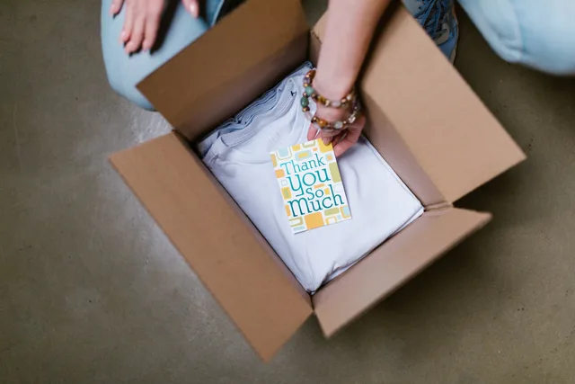 A person packing a white t-shirt with a thank you card into a cardboard box. DTC unit economics